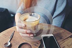 A woman`s hand holding a cup of coffee with smart phone on wooden table in cafe