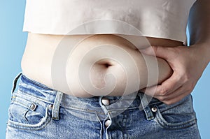 Woman`s hand holding excessive belly fat on blue background, concept of weight loss