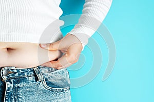 Woman`s hand holding excessive belly fat on blue background, overweight concept