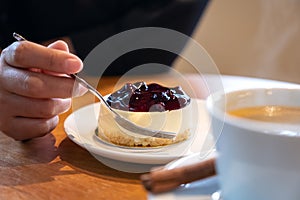 Woman`s hand holding a fork to cut a piece of blueberry cheese cake to eat with coffee cup on wooden table