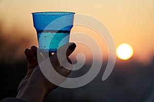 Woman`s hand holding glass against setting sun with blurry skyscrapers in cityscape background