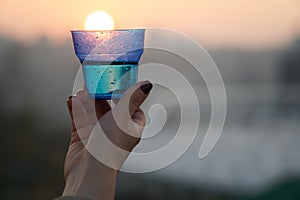 Woman`s hand holding glass against setting sun with blurry skyscrapers in cityscape background