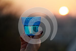 Woman`s hand holding glass against setting sun with blurry skyscrapers in cityscape background