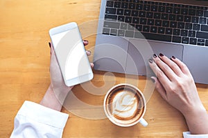 Top view mockup image of a woman`s hand holding mobile phone with blank white desktop screen while using laptop with coffee cup