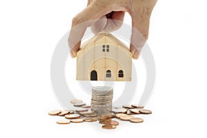 A woman`s hand is holding a model wooden house on stack of coins on white background