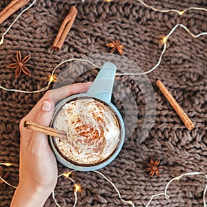 Woman`s hand holding mug of coffee, cocoa or hot chocolate with whipped cream and cinnamon on scarf with pumpkin, leaves