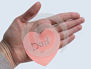Woman`s hand holding pink heart-shaped sticky note with hand written word `Dad` on it on white background. Concept of love, minima
