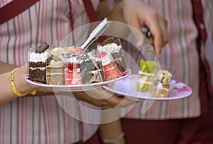 Woman`s hand holding plate of various mini cakes while choosing for takeout at bakery store in outdoor market