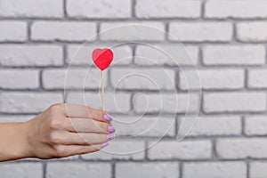Woman`s hand holding red heart shape on stick. White brick wall baskground. Valentine concept.