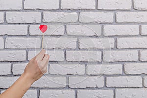 Woman`s hand holding red heart shape on stick. White brick wall baskground. Valentine concept.