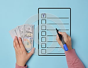 A woman's hand holding a stack of American dollars and filling out a list of purchases or expenses