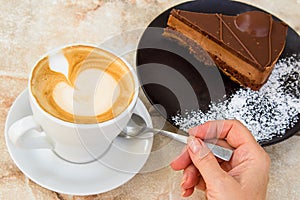 Woman`s hand holding a teaspoon and cup of coffee latte and plate with piece of cake.