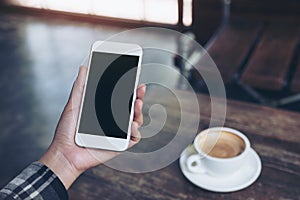 A woman`s hand holding white mobile phone with blank black desktop screen with coffee cup on wooden table in cafe