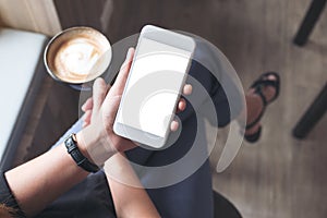 A woman`s hand holding white mobile phone with blank desktop screen while drinking coffee in cafe