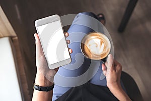 A woman`s hand holding white mobile phone with blank desktop screen while drinking coffee in cafe