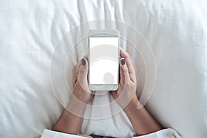 A woman`s hand holding white mobile phone with blank desktop screen on pillow and sitting in the bed