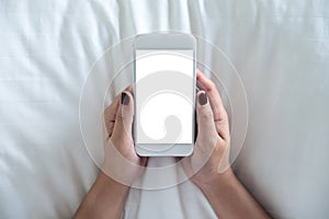 A woman`s hand holding white mobile phone with blank desktop screen sitting in the white bed