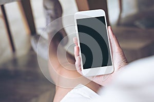 Woman`s hand holding white mobile phone with blank screen on thigh with wooden floor background in modern