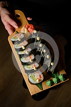 Woman's hand holds appetizing sushi roll futomaki with avocado and crab meat on wooden serving board on black