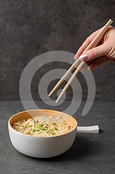 Woman`s hand holds bamboo sticks with noodles in a white bowl bowl on a dark slate stone background