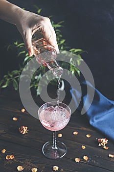 Woman`s hand holds a bottle and pouring champagne in elegant wineglass on dark wooden table surface with dry roses