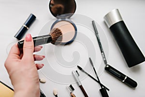 The woman`s hand holds a brush on light white background various set makeup products: brushes, eyeshadow, powder, mascara.
