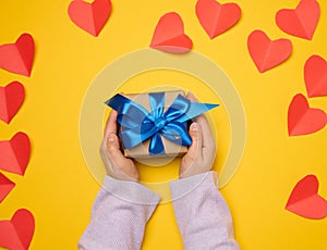 A woman's hand holds a gift box wrapped in a blue silk ribbon on a yellow background