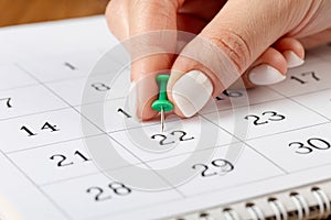 A female hand with a beautiful manicure holds a green pushpin to mark the date on the calendar 22 day