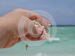 A woman& x27;s hand holds a handful of fine white sand against the background of ocean waves on a sunny summer day on a