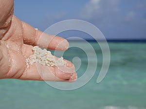 A woman& x27;s hand holds a handful of fine white sand against the background of ocean waves on a sunny summer day on a