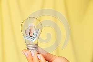 A woman& x27;s hand holds a light bulb and the second hand gives dollars from a light bulb on a yellow background, income