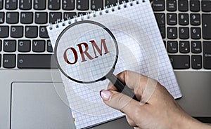 A woman`s hand holds a magnifying glass over the acronym ORM against the background of a laptop