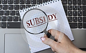 A woman`s hand holds a magnifying glass over the word SUBSIDY against the background of a laptop