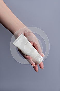 Woman& x27;s hand holds a mockup of white tube of cream. Unlabeled packaging for cosmetics on gray background. Concept of