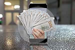 A woman`s hand holds out hundred-dollar bills from a mobile phone, a blurry urban background, raindrops. Concept: transfer money t