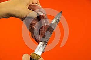 A woman`s hand holds a piece of raw red meat in her hand, in the other hand a large sharp knife cuts the pork. Cooking meat on a