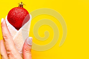 Woman`s hand holds red Christmas ball in paper cone over bright yellow background. Mnimal concept, top view. Xmas or New Year gree