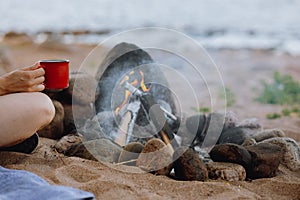 a woman& x27;s hand holds a red cup of coffee or tea against the background of a campfire on a hike. hiking and outdoor