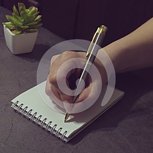 A woman`s hand holds a silver pen and writes in a notebook on the table. The concept of Business, freelancing, planning