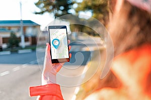 A woman`s hand holds a smartphone with an online map that has a blue geolocation icon. A blurred street is visible in the