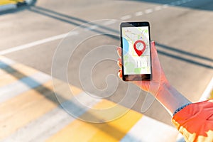 A woman`s hand holds a smartphone with an online map that has a red geolocation icon. In the background, a pedestrian crossing in