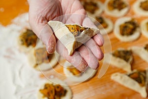 A woman& x27;s hand holds an unbaked amana ear. Sweet gomentashi cookies are on the table in flour next to dried fruits