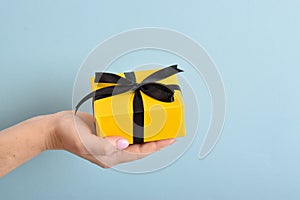 A woman`s hand holds a yellow gift box with a black ribbon on a blue background