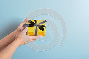 A woman`s hand holds a yellow gift box with a black ribbon on a blue background