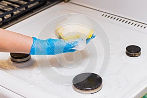 Woman`s hand in a household glove washing sponge with foam a dirty gas stove and burners on kitchen, close up