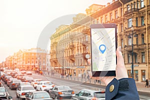 A woman& x27;s hand in a jacket holds a smartphone with an online map app.In the background is a city street with a traffic jam