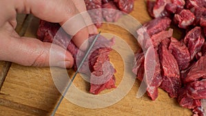 A woman`s hand and a knife cut beef into pieces on a chopping board