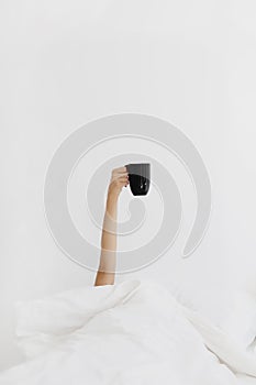 Woman& x27;s hand laying in bed and holding mug with coffee.