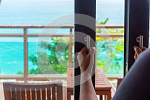 Woman`s hand opening a glass door of hotel room to the ocean view balcony