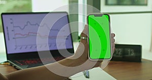 A woman's hand phone with green mock-up screen chroma key holds a smartphone of the workspace background. Stock